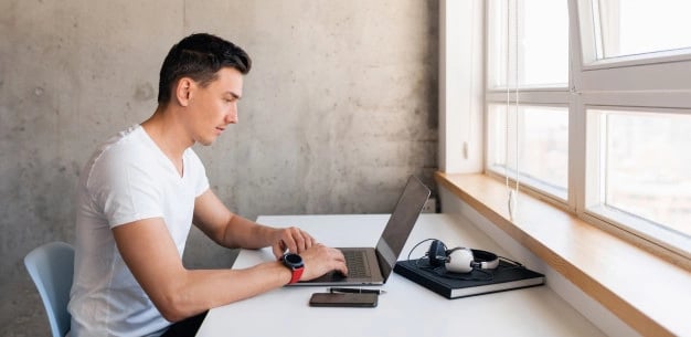 joven-guapo-sonriente-traje-casual-sentado-mesa-trabajando-computadora-portatil-quedandose-solo-casa_285396-1830